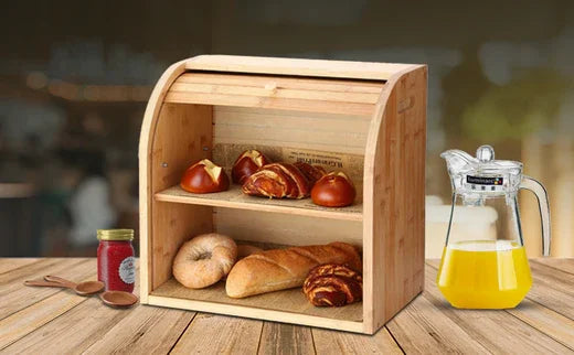 Wooden bread box with assorted fresh bread and croissants on kitchen countertop, jar of jam, and glass jug of orange juice