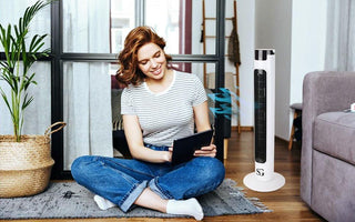 Young woman sitting on rug using tablet with 38 inch white tower fan blowing air in modern living room