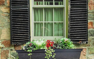 Stone wall window with black shutters and a black woven planter box holding green and red flowers with trailing vines